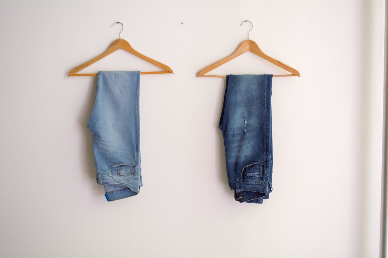 Two pairs of blue denim jeans neatly hanging on wooden hangers against a plain wall.